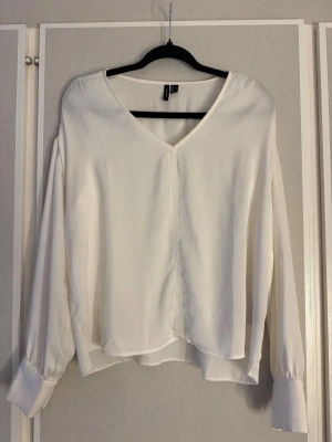 Vit blus med v-ringning  - Stilren vit blus från Vero Moda med v-ringning och långa ärmar. Blusen har breda manschetter och en lös, flowy passform som ger en avslappnad men chic look. .