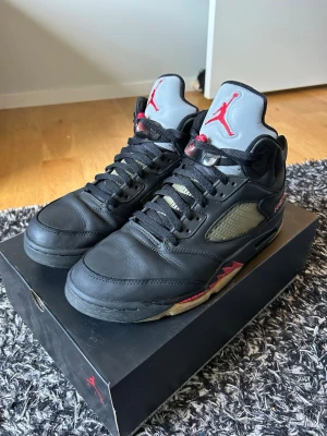 Air Jordan 5 Retro Black/Red Gortex - Säljer ett par Air Jordan 5 Retro gortex sneakers i svart läder med grå och röda detaljer. Skorna har en ikonisk siluett med meshpaneler på sidorna, svart snörning och en grå plös med röd Jumpman-logga. Sulan är svart med röda och beige inslag. Perfekta för dig som gillar streetwear och klassiska sneakers. Syns att de används, har rengjort dem och även lagt på ett tunt lager svart rengörings färg. 