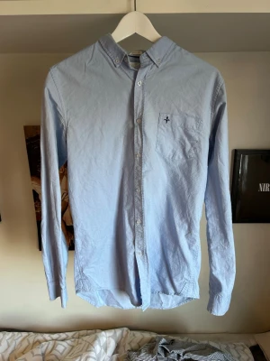 Ljusblå skjorta med knappkrage - Snygg ljusblå skjorta med knappkrage och bröstficka med broderad detalj. Skjortan har långa ärmar och knäppning framtill. Perfekt för en stilren look.