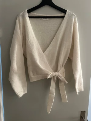 Krämvit omlottstickad tröja Even&Odd - Snygg krämvit stickad omlott-tröja från Even&Odd med knytband i midjan och djup v-ringning. Tröjan har långa ärmar och är perfekt för lager-på-lager-stil. Även jätte mjukt och skönt material🥰