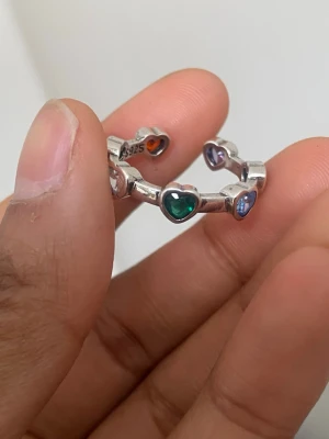s925 sterling Silverring med färgglada hjärtan - Snygg silverring i äkta 925 silver pläterat med hjärtformade stenar i olika färger runt hela ringen. Stenarna är i nyanser av grönt, blått, lila, rött och orange. Perfekt för dig som gillar färg och unika detaljer. 
