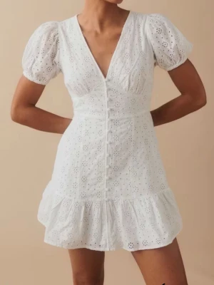 Studentklänning - Superfin vit kort klänning med broderade detaljer, puffärmar och v-ringning. Klänningen har knappar framtill och volang nertill för en söt och somrig look. Perfekt för varma dagar och midsommar. Även perfekt för studenten. Klänningen är från Gina