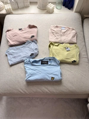 Fem t-shirts från Nike och Lyle & Scott - Fem stilrena t-shirts i olika pastellfärger från Nike och Lyle & Scott. Färgerna är ljusrosa, beige, ljusblå, gul och blå. Alla har rund hals och korta ärmar, med diskreta loggor på bröstet. Perfekta för en avslappnad och trendig look.