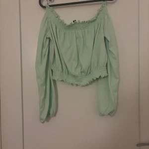 Mintgrön offshoulder blus - Superfin mintgrön blus med offshoulder och långa puffärmar. Smockad resår vid halsringning och midja ger en söt och trendig look. Perfekt att styla med jeans eller kjol för en fräsch vibe.