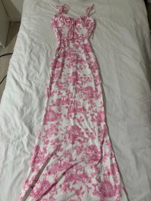 Vit och rosa långklänning med snörning - Långklänning i vit och rosa med snörning framtill och axelband som knyts. Klänningen har ett blommigt mönster och figurnära passform. Perfekt för sommarens alla tillfällen och ger en romantisk vibe. Materialet känns lätt och bekvämt. Aldrig använt🌸🌺