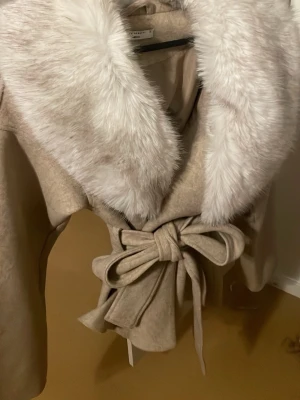 Beige kappa med fuskpäls och knytskärp - Snygg beige kappa från Gina med bred vit fuskpäls på kragen och ett elegant knytskärp i midjan. Kappan har en klassisk och trendig design, perfekt för att hålla sig varm och stilren under kyliga dagar. Materialet är mjukt och känns lyxigt. priset kan såklart diskuteras❤️