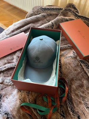 Loro Piana keps - Snygg  blå/grå keps från Loro Piana med broderad logga framtill. Klassisk böjd skärm och justerbar passform. Kepsen är tillverkad i ett mjukt och exklusivt material som ger en lyxig känsla. Perfekt accessoar för en stilren look. Finns en liten defekt som syns på sista bilden, inget man lägger märke till eller tänker på, syns nästan inte alls. Annars är den i toppenskick!