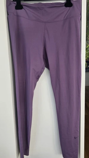 Lila leggings från Nike - Säljer ett par lila träningstights från Nike med hög midja och diskret logga vid benet. Tightsen är i ett stretchigt material som passar perfekt för gym, yoga eller löpning. Snygg passform och bekväma att röra sig i.har några småskador på bakre sömn i grenen men påverkar inte funktionen, använda men i bra skick 