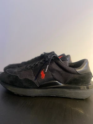 Svarta sneakers från Polo Ralph Lauren - Stilrena svarta sneakers från Polo Ralph Lauren med röd broderad logga på sidan. Skorna har en mix av mocka och textilmaterial, svarta snören och en chunky sula. Perfekta för dig som gillar klassisk men sportig stil.
