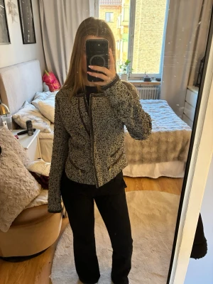 Gråmelerad cardiganjacka med gulddetaljer - Säljer en gråmelerad cardiganjacka med svarta och guldfärgade detaljer längs kanterna och vid fickorna. Jackan har en rak passform, långa ärmar och två fickor med guldfärgade dragkedjor. Perfekt för att lyfta en outfit.