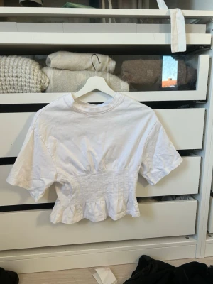 Vit croppad topp med markerad midja - Snygg vit croppad topp med korta ärmar och markerad midja som ger en feminin siluett. Toppen har en lätt rynkad effekt nedtill och är tillverkad i mjuk bomull. Perfekt att styla med jeans eller kjol för en trendig look.