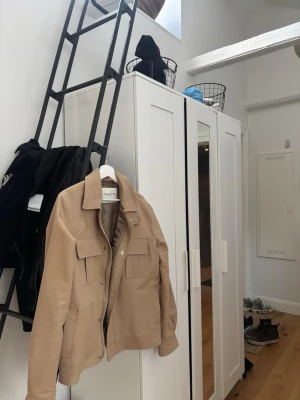 Beige jacka från These Glory Days - Stilren beige jacka från These Glory Days med klassisk krage, två stora bröstfickor med lock och knappar samt dragkedja framtill. Jackan har en rak passform och är tillverkad i ett mjukt bomullsmaterial. Perfekt för dig som gillar minimalistisk stil.sitter som en medium i gott skick 