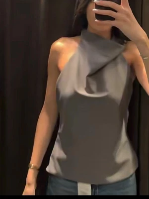 Grå axelbandslös halterneck topp ZARA - Snygg grå axelbandslös halterneck topp med öppen rygg och draperad halsringning. Perfekt till jeans eller kjol för en trendig look. Tunn och mjuk känsla, troligtvis i polyester eller liknande material.
