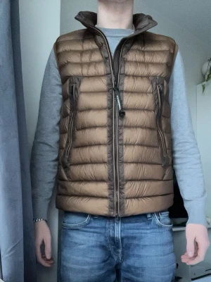 Brun dunväst med märket av c.p Company  - Snygg brun dunväst med hög krage och två vertikala dragkedjefickor framtill. Västen har quiltad design med horisontella sömmar och stängs med dragkedja. Perfekt att slänga över en hoodie eller tröja för extra värme och stil.