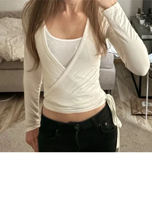 Vit omlottopp med knyt i sidan från Nelly  - Säljer en vit omlottopp med långa ärmar och knyt i sidan. Toppen har en croppad passform och är tillverkad i mjukt bomullsmaterial. Pris kan diskuteras 