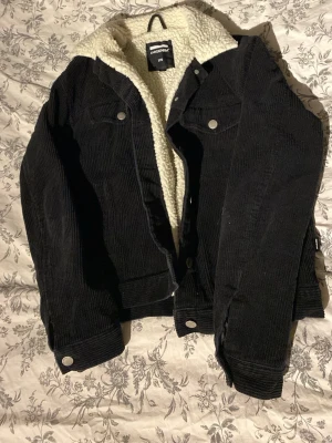 Svart manchesterjacka med teddyfoder - Svart manchesterjacka från Monki i storlek XS med mysigt vitt teddyfoder. Jackan har knappar framtill, två bröstfickor med lock och klassisk krage. Perfekt för lager-på-lager och en cool streetstil.