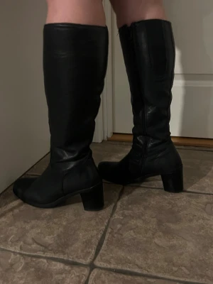 Svarta knähöga boots med klack - Snygga svarta knähöga stövlar i slätt läderimitation med bred klack och rundad tå. Stövlarna har dragkedja på insidan och en stilren design som passar till många outfits. Perfekta för dig som vill ha en klassisk och trendig look.