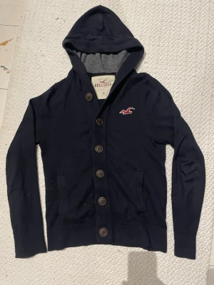 Marinblå hoodie från Hollister - Snygg marinblå hoodie från Hollister med knappar framtill och klassisk logga på bröstet. Hoodien har två fickor, ribbade muddar och en mjuk insida. Perfekt för dig som gillar en avslappnad och stilren look.