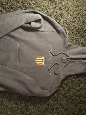 Mörkgrå hoodie från GANT - Mörkgrå hoodie från GANT med broderad logga och amerikanskinspirerat sköldmärke på bröstet. Klassisk känguruficka framtill och justerbar dragsko i huvan. Mjuk bomullsblandning som är perfekt för en avslappnad stil.