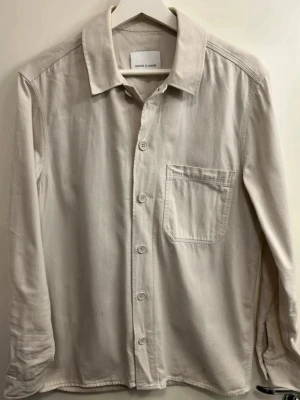 Beige overshirt från Samsøe Samsøe - Stilren beige overshirt från Samsøe Samsøe med klassisk krage, knappstängning och en bröstficka. Overshirten har raka linjer och är tillverkad i ett mjukt bomullsmaterial. Perfekt att bära öppen eller stängd över en t-shirt.
