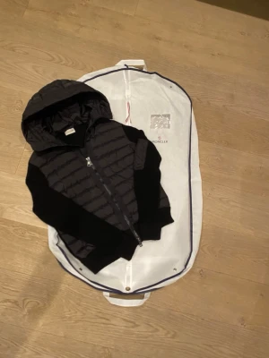 En svart moncler cardigan i perfekt skick perfekt för dig som vill uppleva strettwere - Snygg svart dunväst från Moncler med stickade svarta ärmar och stor huva. Västen har dragkedja framtill och klassisk Moncler-logga på insidan. Perfekt för lager-på-lager och streetstyle.
