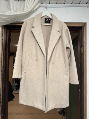 Beige ullkappa från Pull & Bear - Stilren beige kappa från pull and bear med klassisk krage och två fickor framtill. Kappan har dubbelknäppning och är tillverkad i en mjuk ullblandning. Perfekt för att hålla värmen under kyliga dagar och ger en clean look till din outfit.