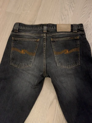 Nudie Jeans  - Säljer ett par svarta jeans från Nudie Jeans med klassisk femficksdesign och snygga orange sömmar på bakfickorna. Jeansen har en rak passform och är tillverkade i denim av bomull. Perfekta för en stilren och avslappnad look.