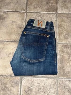 Nudie jeans  - Säljer ett par Nudie Jeans Skinny Lin i mörkblå tvätt med klassiska orange sömmar. Modellen är skinny fit med normal midja och tillverkad i stretchig denim. Snygga detaljer och ikonisk Nudie-logga på bakfickan. Midja 31cm, längd 94cm. JAG SKICKAR EJ FLER BILDER NÄR JAG BÄR JEANSEN! för storleksguide hänvisar jag till måtten och för bättre inblick i passformen rekommenderar jag att googla på modellnamnet. kolla måtten noggrant :) s4 