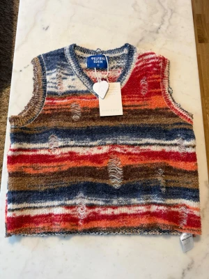 Randig stickad väst från Elfric Eden - Färgstark stickad väst från Elfric Eden med breda ränder i blått, rött, brunt och vitt. Västen har en avslappnad passform, V-ringning och slitna detaljer för en edgy look. Perfekt för dig som vill sticka ut med din stil.