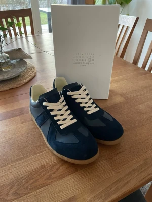 Maison Margiela navy sneakers - Snygga sneakers från Maison Margiela i mörkblå mocka och läder med beige snörning och klassisk gummisula. Låg modell med stilren design och diskreta detaljer. Perfekta för dig som gillar minimalistisk och trendig stil.