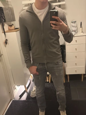 Grå ziptröja från H&M - Snygg grå ziptröja från H&M med hög krage och dragkedja hela vägen. Tröjan är tunnstickad och har ribbade muddar vid ärmslut och nederkant. Perfekt att slänga över en t-shirt för en clean look.