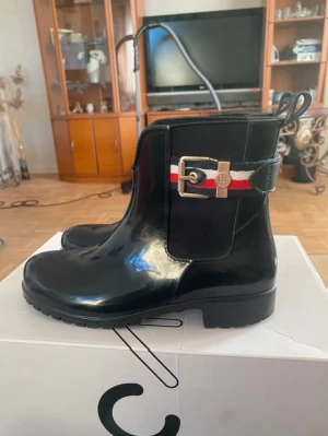 Gummistövlar, Tommy Hilfiger. - Säljer vidare mina snygga gummistövlar från Tommy Hilfiger med dekorativt spänne och röd-vit-blått band på sidan. Stövlarna har låg klack, rund tå och dragflik baktill. Perfekta för regniga dagar/vår och har en klassisk, stilren look. Storlek, 37. 🌸