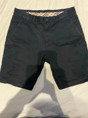 Svarta shorts från Burberry - Snygga svarta shorts från Burberry med klassiskt rutigt foder i midjan. Shortsen har normal passform, bälteshällor, sidofickor och bakfickor med knapp. Perfekta för sommaren och stilrena till alla tillfällen.