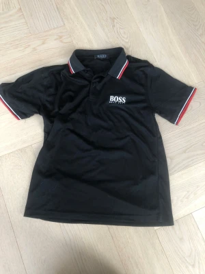 Svart pikétröja från Hugo Boss - Klassisk svart pikétröja från Hugo Boss med vit logga på bröstet. Tröjan har krage och knappar framtill samt röda och vita detaljer på krage och ärmslut. Perfekt för en stilren och sportig look.