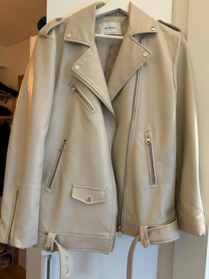 Beige skinnjacka från Gina Tricot - Jättesnygg beige skinnjacka från Strandivarius med fina detaljer. Jackan har flera silverfärgade dragkedjor och bälte i midjan. Perfekt för dig som vill ha en stilren och trendig look. Nacken på jackan är något sliten, se näst sista bild men syns ej märkbart när jackan bärs samt en liten fläck på armen de sista bild annars fint skick🤍🤍🤍