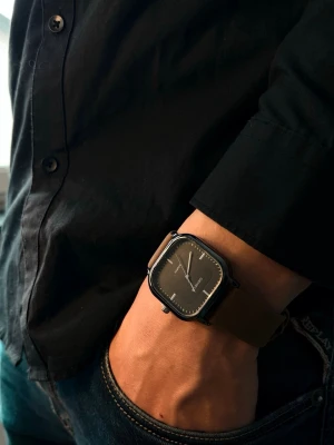 Svart kvadratisk klocka  - Stilren klocka med svart kvadratisk urtavla och minimalistisk design. Klockan har en brun rem i skinn och silverfärgade visare. Perfekt accessoar för dig som gillar modern och enkel stil.