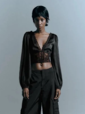 Svart croppad blus med spetsdetaljer - Snygg svart croppad blus med djup v-ringning och spetsdetaljer längs kanten. Blusen har långa, lite puffiga ärmar och transparent tyg över magen. Perfekt för dig som vill ha en trendig och edgy look.