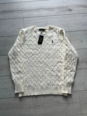 Vit kabelstickad tröja Polo Ralph Lauren - Kabelstickad vit tröja från Polo Ralph Lauren med rund halsringning och broderad logga på bröstet. Tröjan har ribbade muddar vid ärmslut och nederkant, och är tillverkad i mjukt material som ger en klassisk och stilren look.