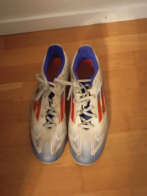 Adidas f50 storlek  - Säljer ett par snygga Adidas fotbollsskor i vitt med röda och blå detaljer. Skorna har klassisk snörning, blå insida och mönstrad ovandel. Perfekta för dig som vill sticka ut på planen med stilrena och sportiga skor.