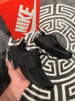Svarta Nike Air Presto sneakers - Säljer ett par helsvarta Nike Air Presto sneakers med mesh-ovandel och plastdetaljer vid snörningen. Skorna har en låg silhuett, bekväm sula och klassisk Nike-logga på sidan. Perfekta för dig som gillar stilrena och sportiga sneakers.