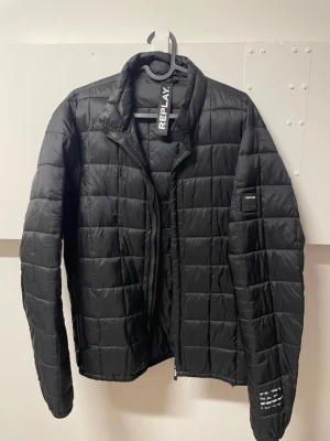 Svart dunjacka från Replay - Snygg svart dunjacka från Replay med quiltat mönster och hög krage. Jackan har dragkedja framtill, Replay-logga på ärmen och en stilren design som passar perfekt till vintern. Lätt och smidig att bära, med modern passform.