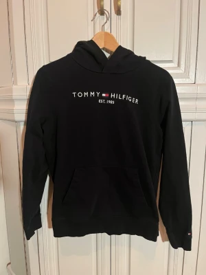 Svart hoodie från Tommy Hilfiger - Svart hoodie från Tommy Hilfiger med vit och röd logga samt texten 'TOMMY HILFIGER EST. 1985' på bröstet. Klassisk känguruficka framtill och huva. Endast provad en gång, glömde skicka tillbaka den