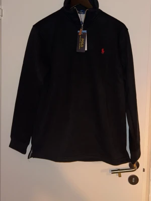 Svart quarter zip från Polo Ralph Lauren - Snygg svart långärmad tröja från Polo Ralph Lauren med halv dragkedja och klassisk röd logga broderad på bröstet. Tillverkad i mjukt material som passar perfekt till en avslappnad stil. Enkel och stilren design. Hejsan alla! Jag har precis fyllt 18 år och har fått en massa presenter. Det är mycket av presenterna som inte passar min stil,  Det är därför jag väljer att sälja detta.  Allt är oanvänt, och prislapparna är kvar Polo Ralph lauren hoodie, Storlek L men passar även för större M pris 899