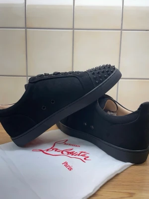 Svarta Christian Louboutin sneakers - Snygga svarta sneakers från Christian Louboutin i mocka med svarta nitar på tån. Skorna har svarta snören, diskret logga på sulan och klassisk låg profil. Perfekta för dig som vill sticka ut med en edgy och exklusiv look.
