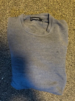 Blå stickad tröja från J.Lindeberg - Snygg blå stickad merino tröja från J.Lindeberg med rund halsringning och diskret logga broderad på bröstet. Tröjan har en klassisk passform och är tillverkad i ett mjukt, tunt material som passar perfekt till lager-på-lager-stil.