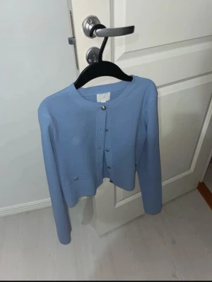 Ljusblå cropped kofta med knappar - Snygg ljusblå cropped kofta med rund halsringning och silverfärgade knappar framtill. Koftan har långa ärmar och en liten ficka med dekorativ knapp. Perfekt att styla över ett linne eller t-shirt.