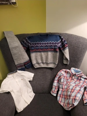 Klädpaket med stickad Tröja Halsduk och Skjortor 3år - Detta är ett klädpaket med en stickad tröja med ett traditionellt mönster i blått, grått och rött. Det finns även ett par grå stickade byxor med matchande mönsterdetaljer. En vit kortärmad skjorta med subtil textur och en röd- och blårutig långärmad skjorta med broderi på bröstet ingår också. Passar ca 3 år barn