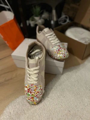 Maison Margiela sneakers paint on - HELT NYA. Unika sneakers från Maison Margiela i beige mocka och läder med färgstänk i rött, gult, grönt och vitt på tån.  Snygga skor som  sticker ut i mängden. De är replika och inte äkta så ni vet, köpte dom nyss från en annan på plick och de är för små, storlek skulle jag säga 42 även fast det står 43.