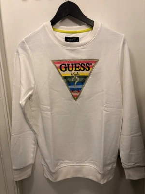 Guess Vit sweatshirt  - Vit sweatshirt från Guess med stort broderat trekantsmärke på bröstet i regnbågsfärger och glittriga detaljer. Klassisk rund halsringning och ribbade muddar. Perfekt för dig som gillar streetstyle och vill sticka ut med en ikonisk logga.