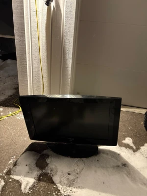 Svart Samsung TV med platt skärm - En svart platt TV från Samsung med bred ram och rund fot. Skärmen är rektangulär och har en stilren design som passar bra i de flesta rum. Perfekt för dig som vill koppla in spelkonsol eller använda som extra skärm.